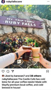 Two hands each hold an iced coffee in front of a stone arch with a sign reading My Trip Thru Ruby Falls